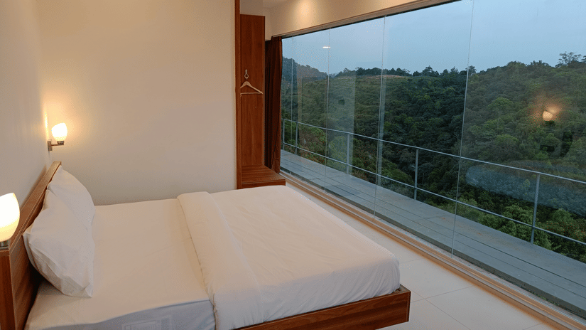 The First Floor Corner Room at The Cliff Edge Coorg Hotel featuring a large bed and floor-to-ceiling glass windows offering panoramic views of the lush green forest.