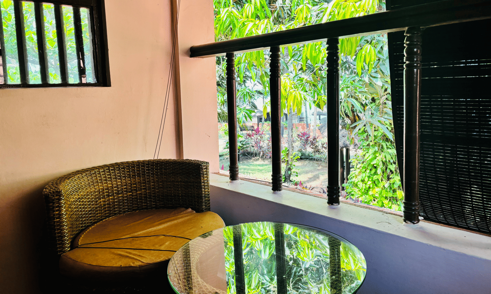 Balcony with round glass-top table and a wicker chair overlooking greenery at Paradise Lagoon Resort, Udupi.