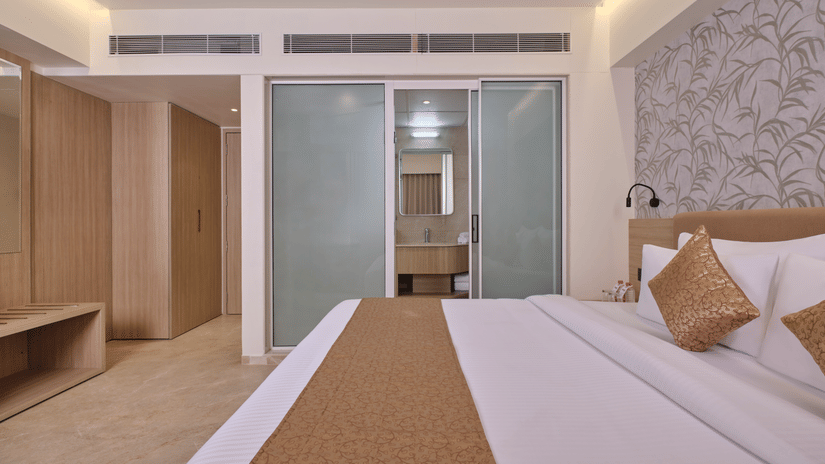 A bedroom with a built-in wooden wardrobe, luggage rack, and bathroom doorway with the bed in the foreground at Biznotel by Pride Motera, Ahmedabad.