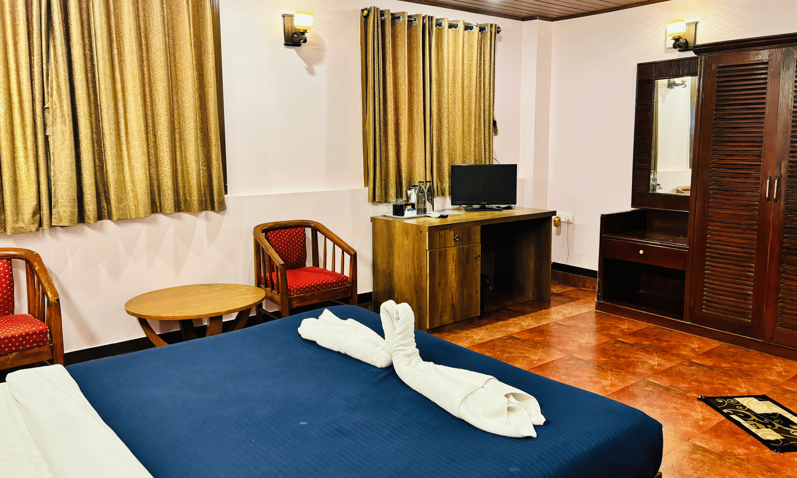 Interior of a standard room at Paradise Lagoon Resort, Udupi, featuring a double bed, wooden furniture including a desk and wardrobe, gold-colored curtains, and a seating area with red cushions.
