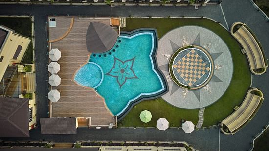 Aerial view of a luxurious hotel pool area with a floral design in the pool, surrounding lounge chairs with umbrellas, and a circular plaza featuring a large chessboard pattern - Voyage The Solitaire Bliss.