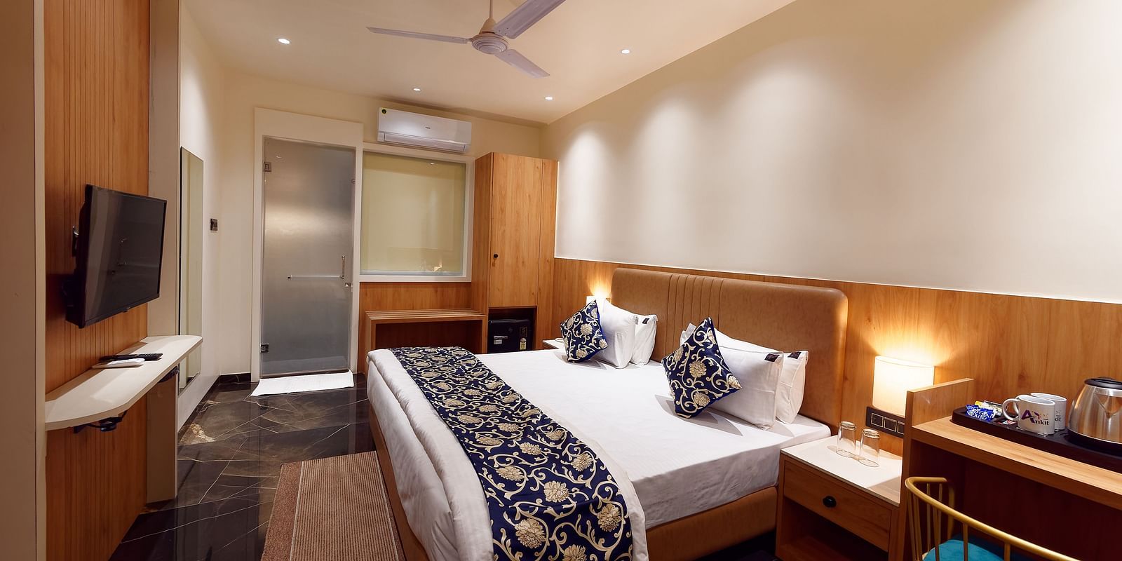 Image of deluxe room with a bed with table and chair on the side of the table and a fan on te ceiling at Ankit Hotel, Jabalpur