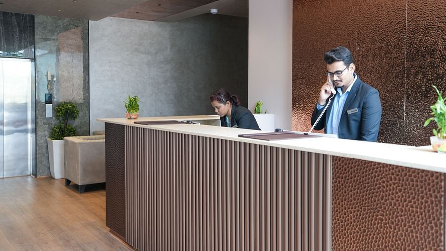 Close-up of reception desk with staff assisting guests, showing copper-toned wall and wooden flooring at TGI Livancia Hillside, JC Nagar, Mysore