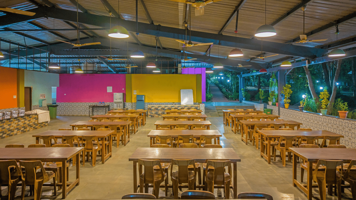 Spacious resort dining hall with rows of wooden tables and benches, vibrant yellow, pink and blue painted walls, ceiling fans and bright overhead lighting.
