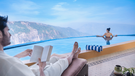 Couple enjoy the Presidential Suite infinity pool at Polo Resort Cherrapunji, one reading on a lounger in a robe while another guest looks out at cliffs and waterfalls.