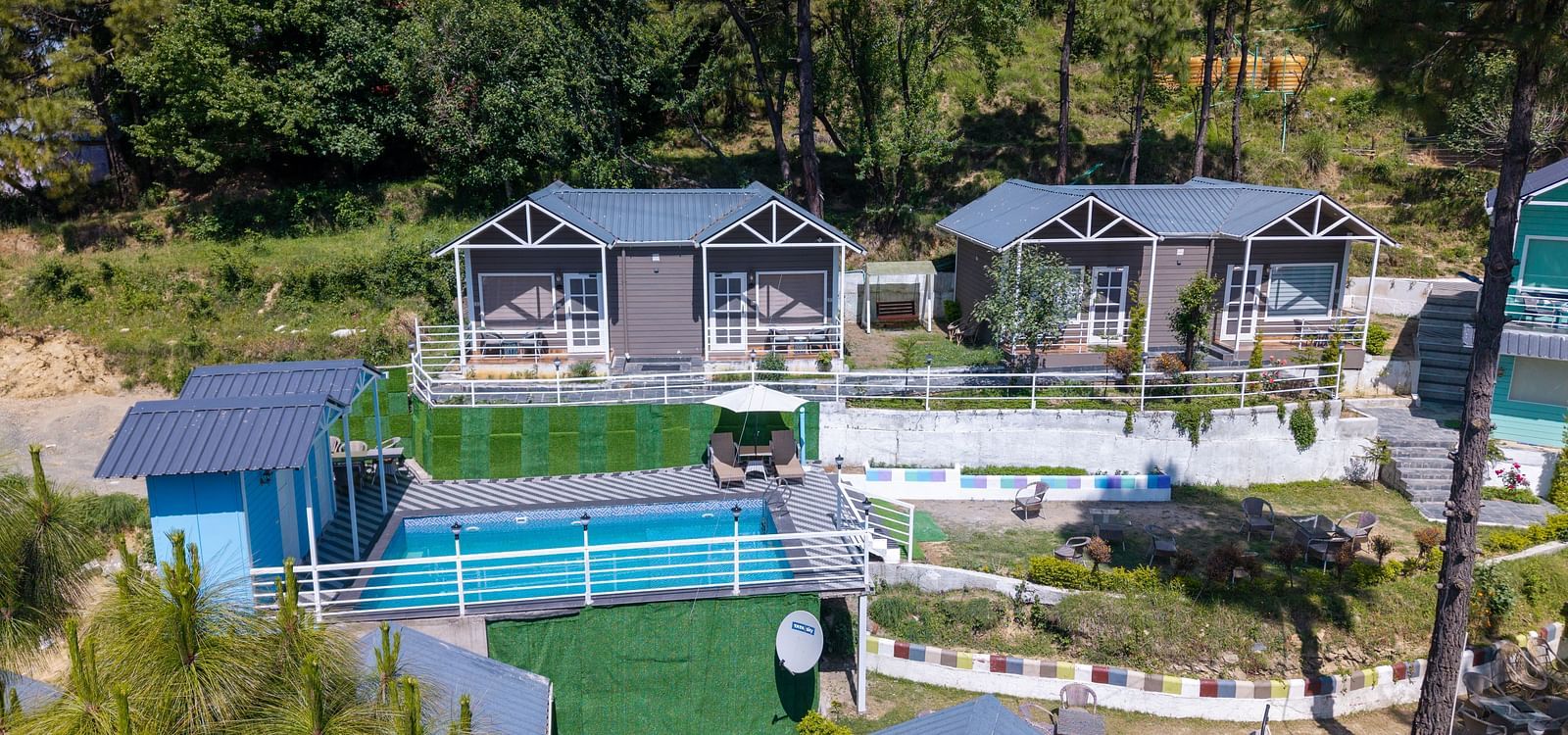 High-angle shot of two modern cabins with large windows, situated on a terraced hillside overlooking a blue swimming pool. Lush green trees surround the property - DLS Nature Trinket Resort, Dalhousie.