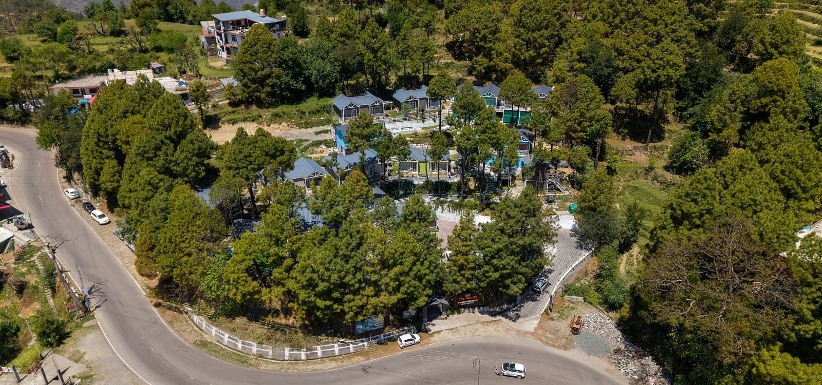 An elevated shot shows a winding road through a hilly landscape with trees and buildings. Cars are visible on the road, and the mix of residential and commercial structures is bathed in daylight under a clear sky - DLS Nature Trinket Resort, Dalhousie.