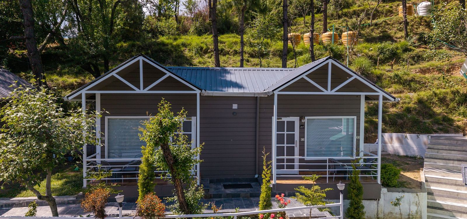 Charming hillside cottage with a twin-gabled roof, large front windows, and a landscaped garden surrounded by trees and greenery - DLS Nature Trinket Resort, Dalhousie.