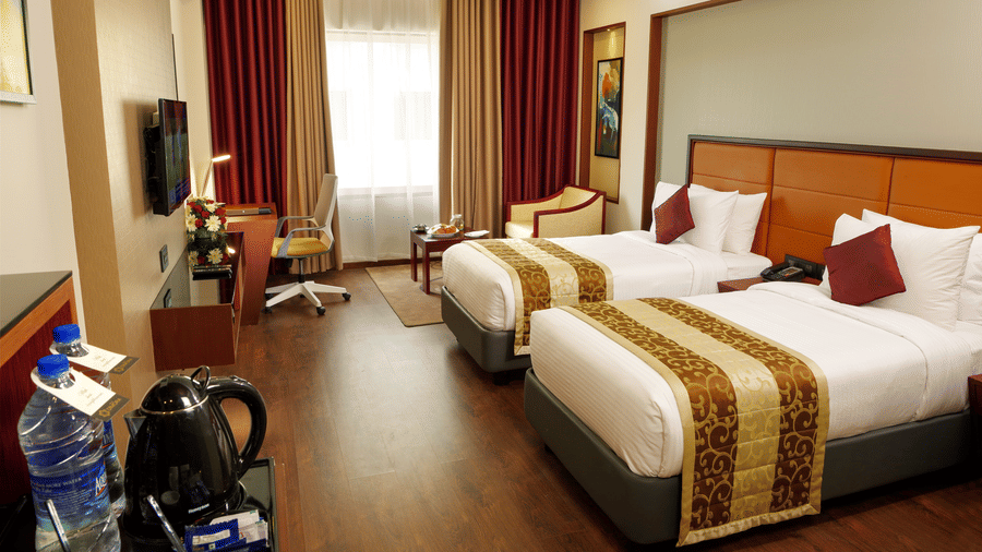 The Standard Room with twin beds at Dimora, Thiruvananthapuram, with twin beds, a flat screen television, glass window with curtains, and wodden flooring.