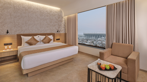 A modern hotel room featuring a king size bed, seating area with a sofa, and a large window overlooking the city skyline at Biznotel by Pride Motera, Ahmedabad.