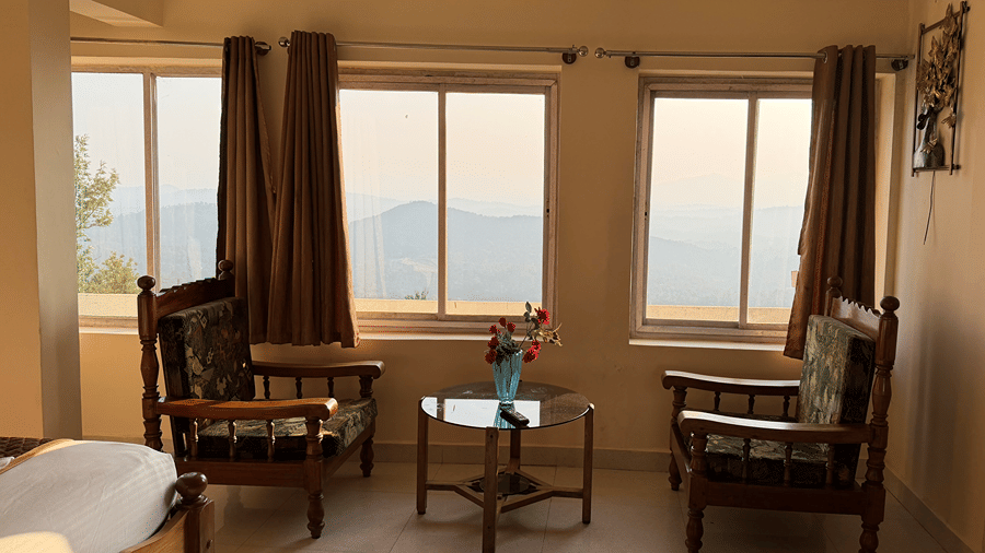 A seating area with two wooden armchairs and a small coffee table positioned next to large windows | Kollanoor Guest House, Coorg