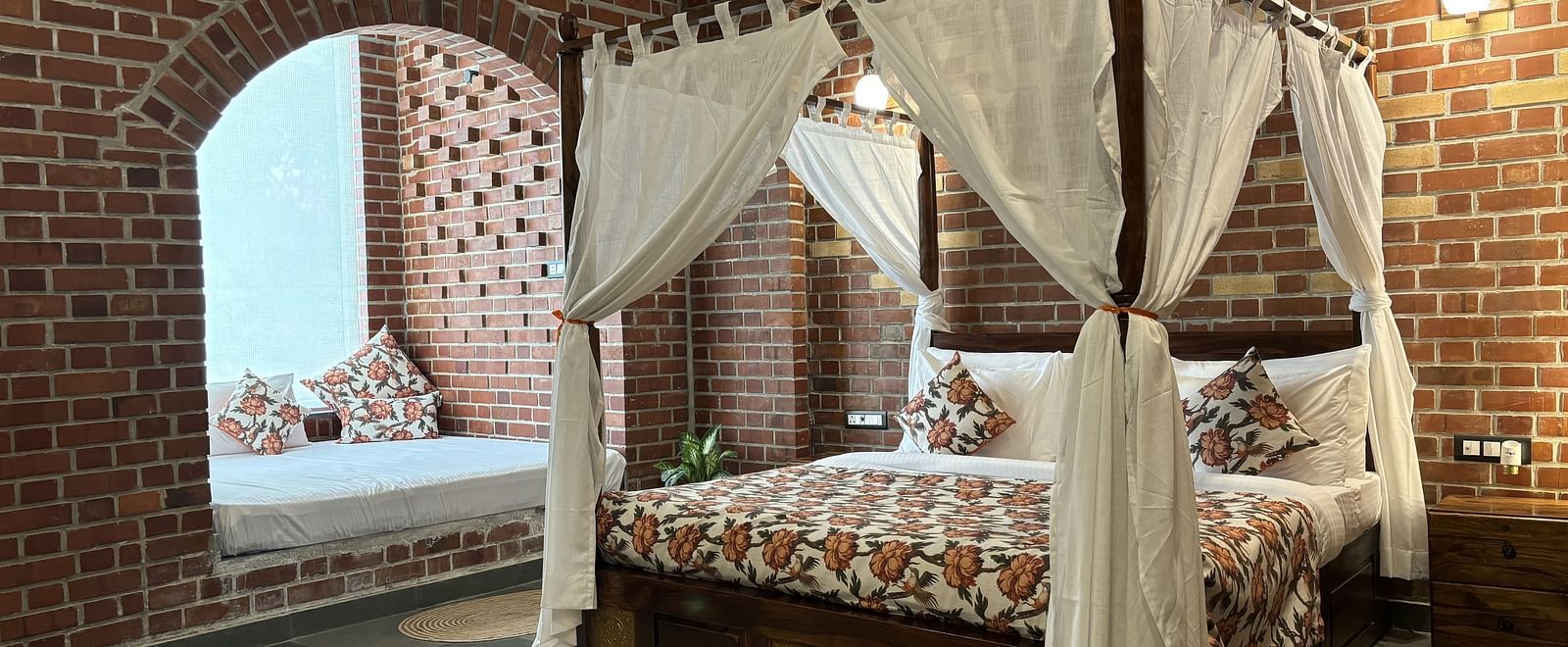 A bedroom at Tisya Farms, Gurgaon with a four-poster bed, white curtains, brick walls, and an arched window.