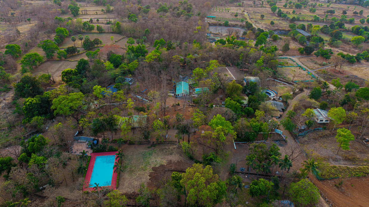 Aerial drone view of Nature Trails Sajan resort surrounded by dense greenery, featuring a blue swimming pool, red-roofed buildings, and vast landscape.