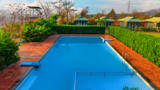 Swimming pool at Nature Trails Durshet surrounded by lush greenery, cottages, and scenic hillside views under a bright blue sky.