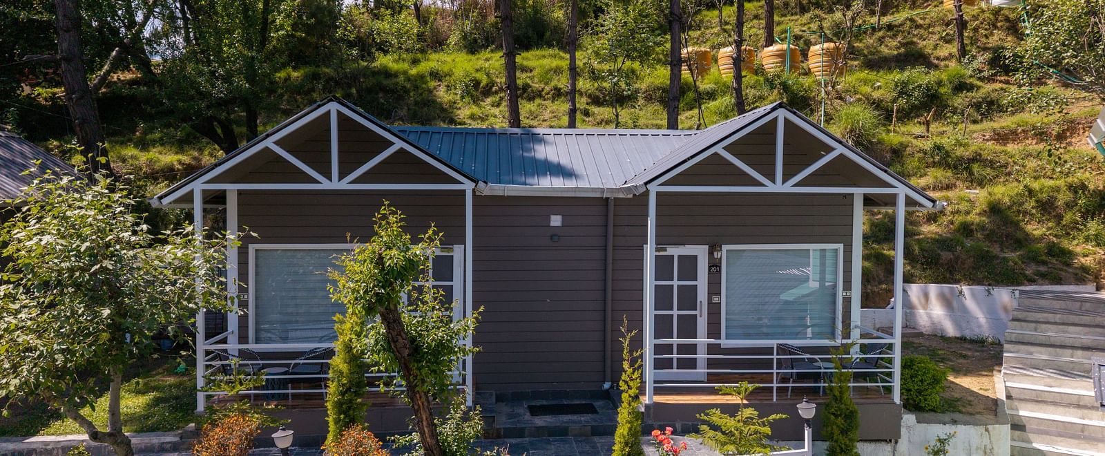 Charming hillside cottage with a twin-gabled roof, large front windows, and a landscaped garden surrounded by trees and greenery - DLS Nature Trinket Resort, Dalhousie.
