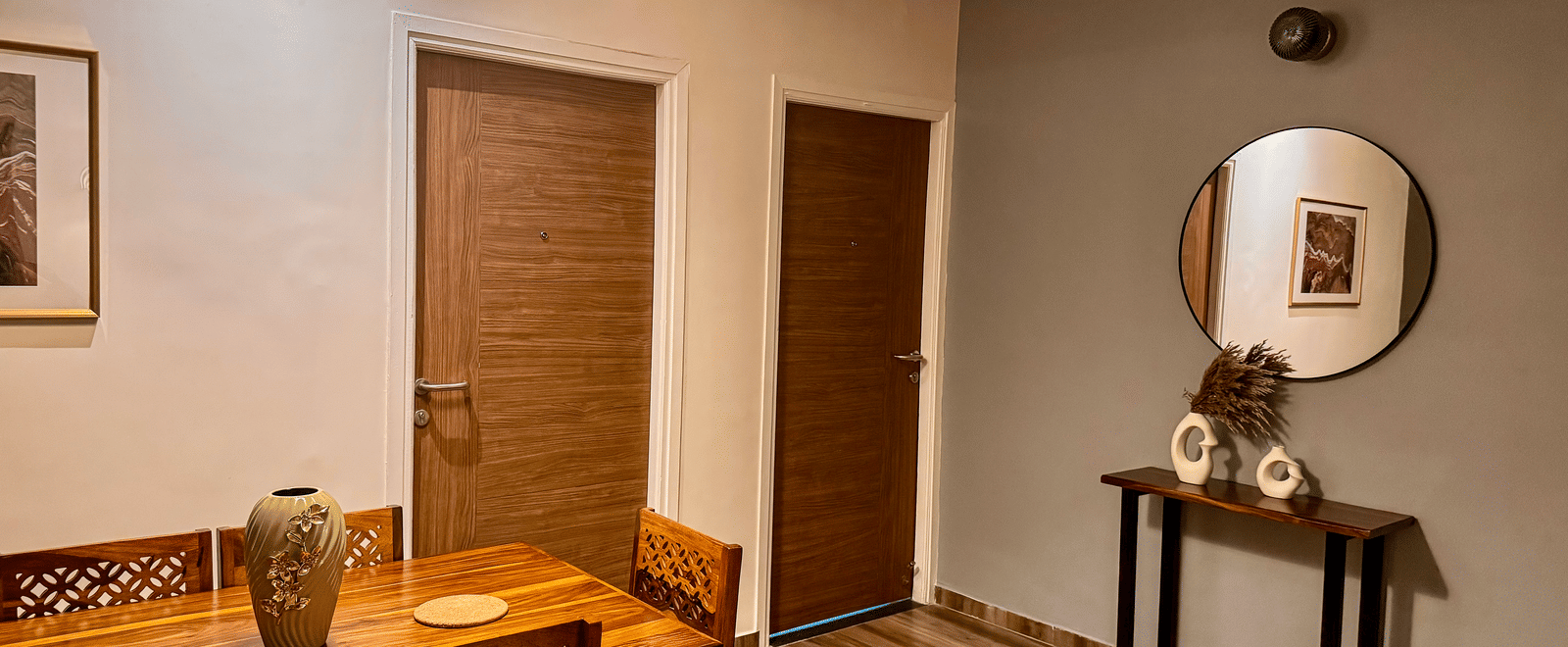 Common area in the Coral 4BHK featuring a wooden dining table, wooden flooring, a picturesque wall set-up with a round mirror, a small table below it, with a vase on it | Kove Smart Stays