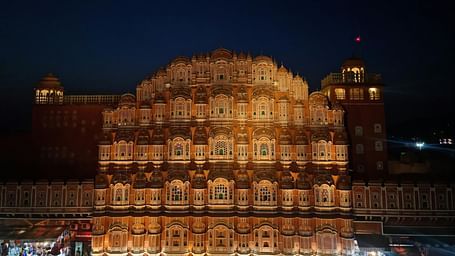 The Hawa Mahal in Jaipur is illuminated at night, showcasing its intricate facade and numerous small windows against a dark blue sky.