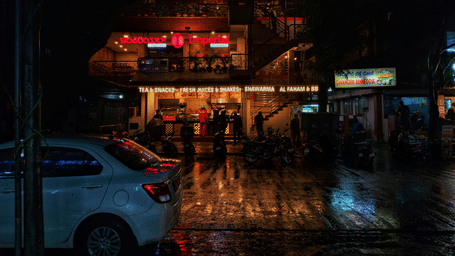 A wet city street at night reflecting neon lights and signage, with a car parked on the side and people visible in the background