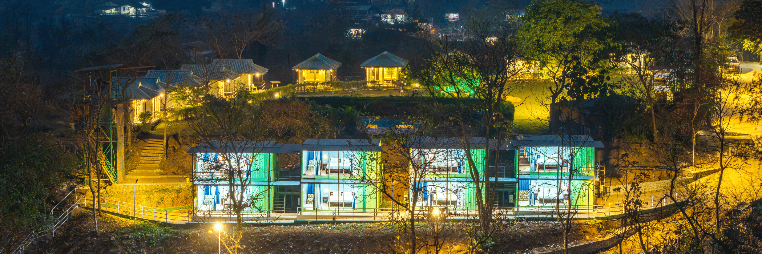 Alt Text: Aerial night view of Nature Trails Durshet resort glowing with warm lights nestled among lush hills and trees.