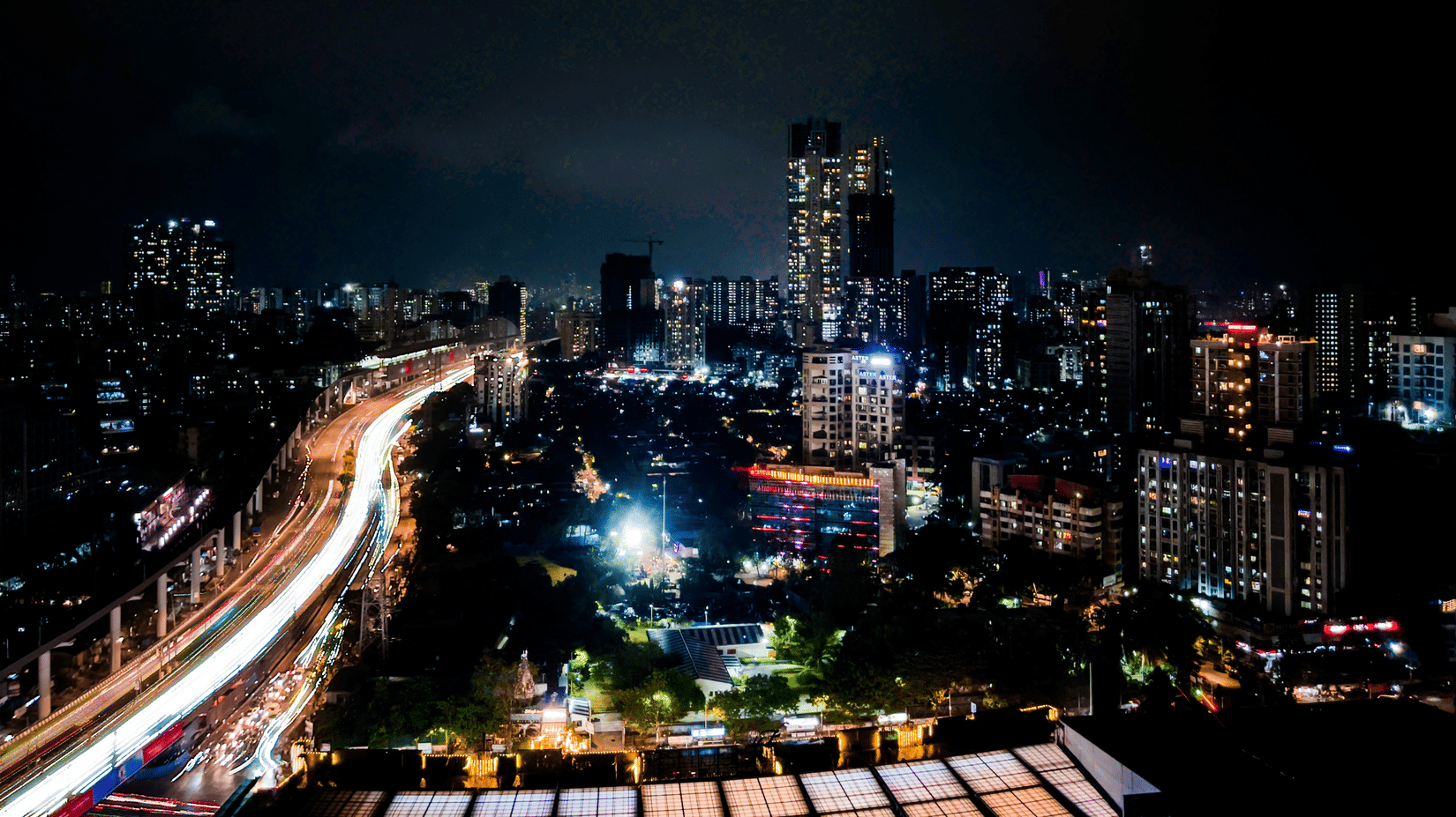Night-time city skyline with illuminated buildings, elevated roads, and light trails stretching across the urban landscape