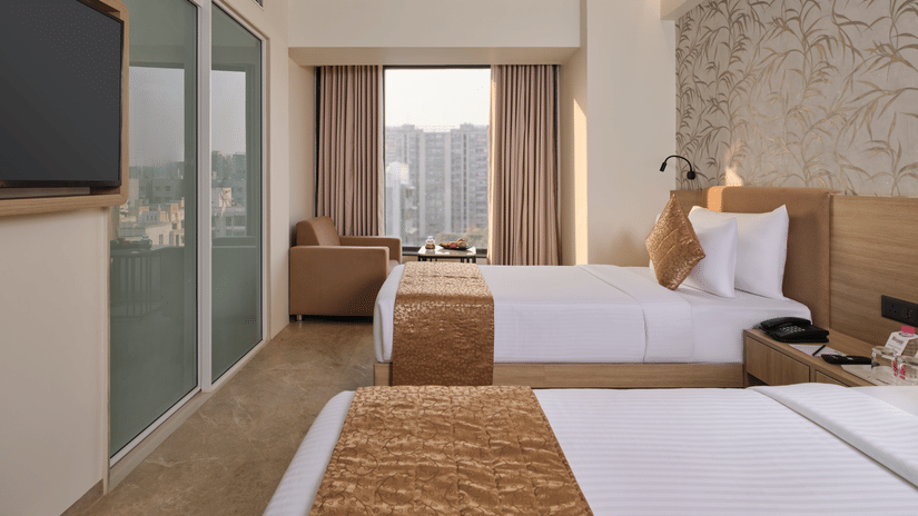 A modern hotel room furnished with twin beds, gold bed runners, and a large window looking out toward city buildings at Biznotel by Pride Motera, Ahmedabad.