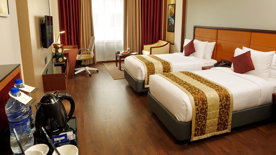 The Standard Room with twin beds at Dimora, Thiruvananthapuram, with twin beds, a flat screen television, glass window with curtains, and wodden flooring.