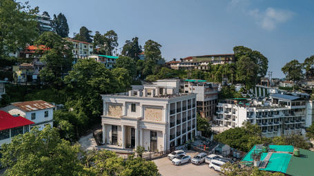 An elevated view shows a  multi-story building surrounded by green hills and other structures under a clear sky.