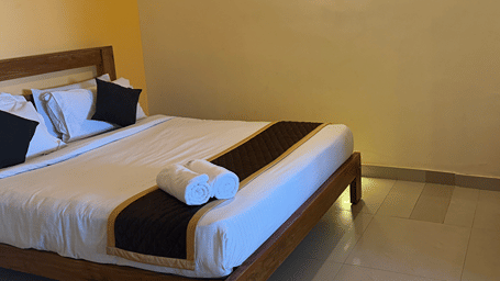 A bedroom with a wooden bed frame, white bedding, and a small side table covered with a patterned red tablecloth | Kollanoor Guest House, Coorg