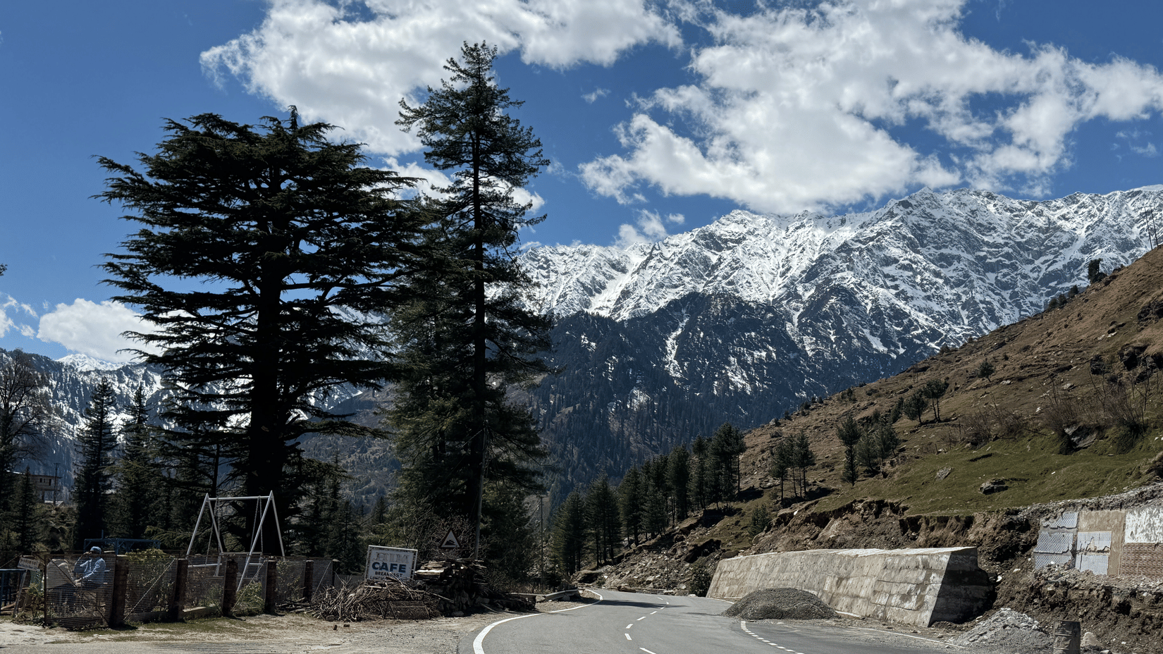 A road curves past trees towards mountains under clouds. Snow covers peaks in the distance. A structure sits by the roadside.