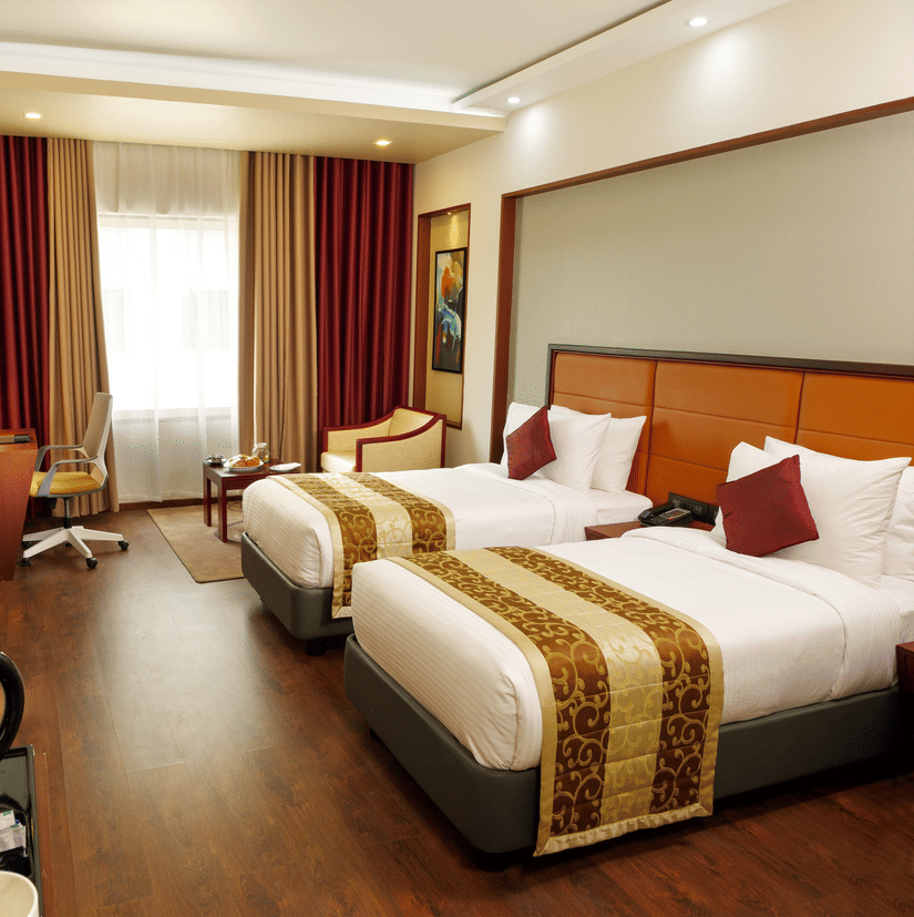 The Standard Room with twin beds at Dimora, Thiruvananthapuram, with twin beds, a flat screen television, glass window with curtains, and wodden flooring.
