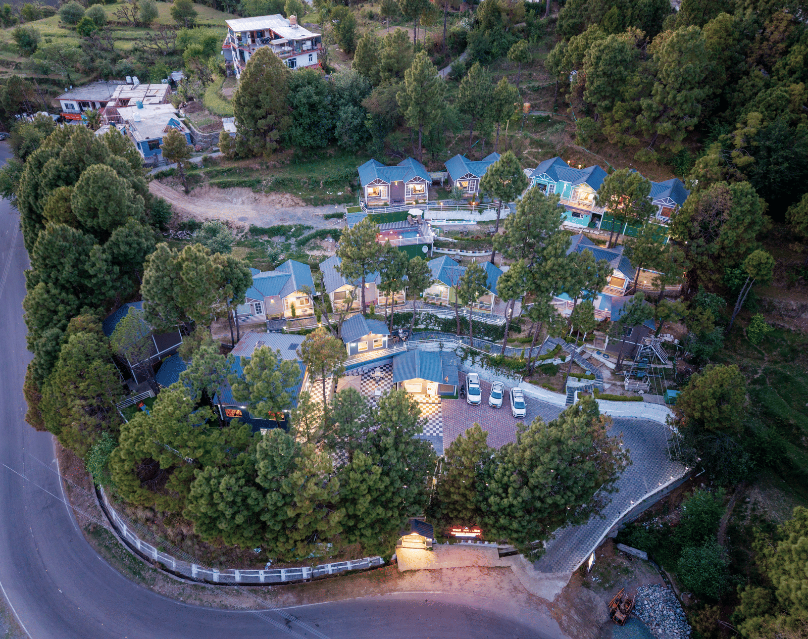 Aerial view of a scenic hillside resort featuring blue-roofed cottages, winding roads, and lush green trees, with nearby rural structures and natural surroundings - DLS Nature Trinket Resort, Dalhousie.