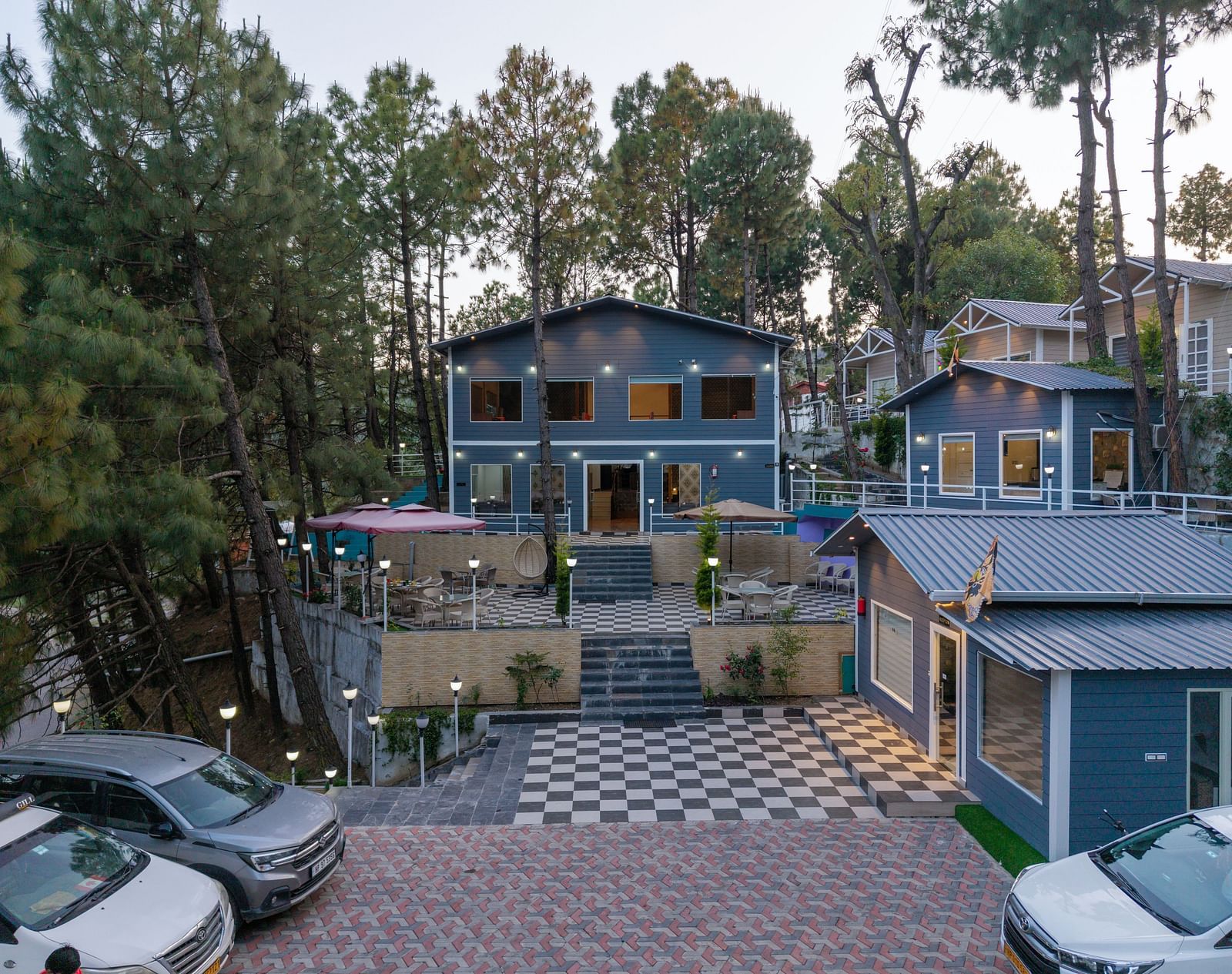 Two modern buildings nestled among tall pine trees with parked cars in the foreground - DLS Nature Trinket Resort, Dalhousie.