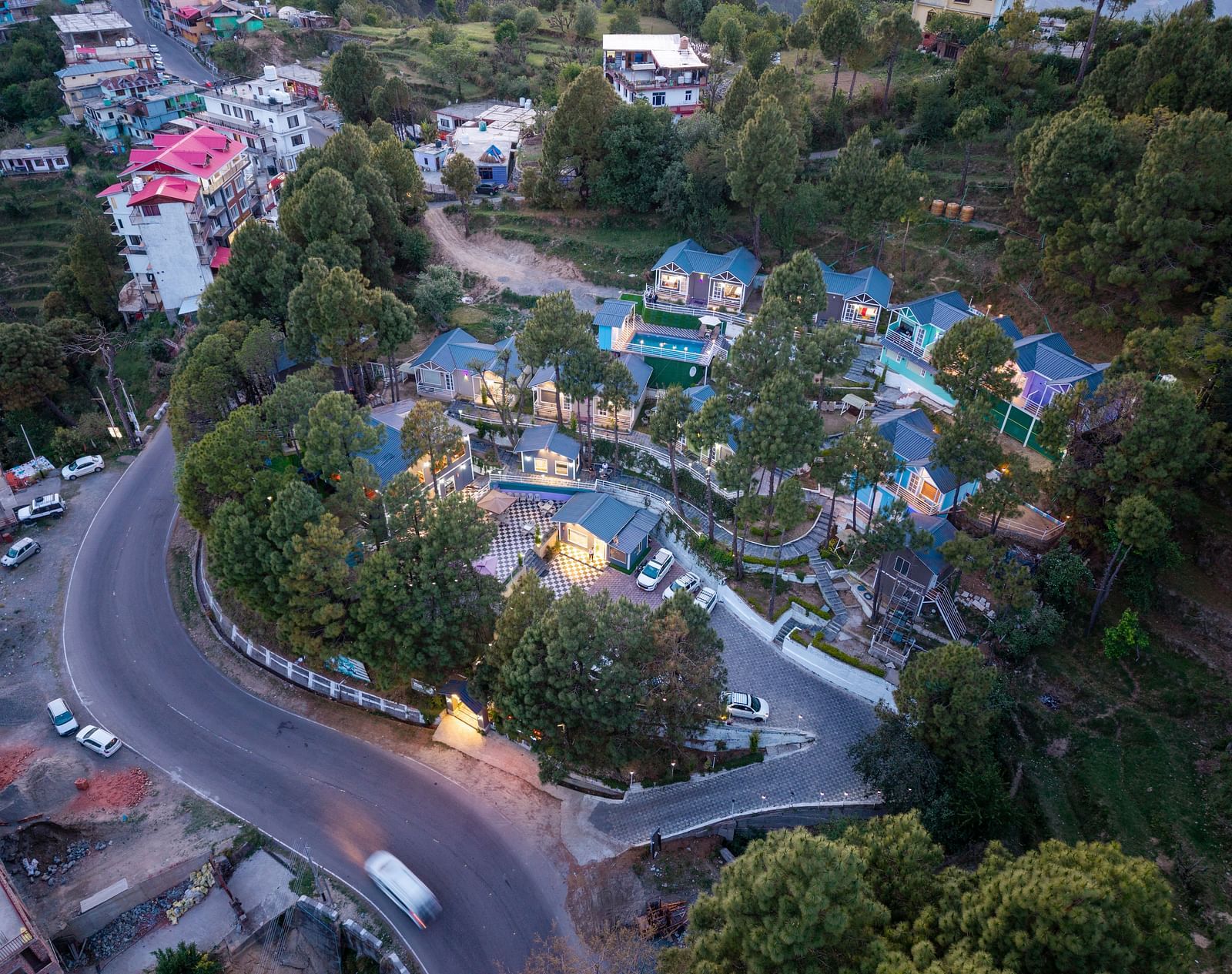 Aerial view of a hillside resort complex with blue-roofed cottages, located alongside a winding road and surrounded by lush greenery and nearby town buildings - DLS Nature Trinket Resort, Dalhousie.