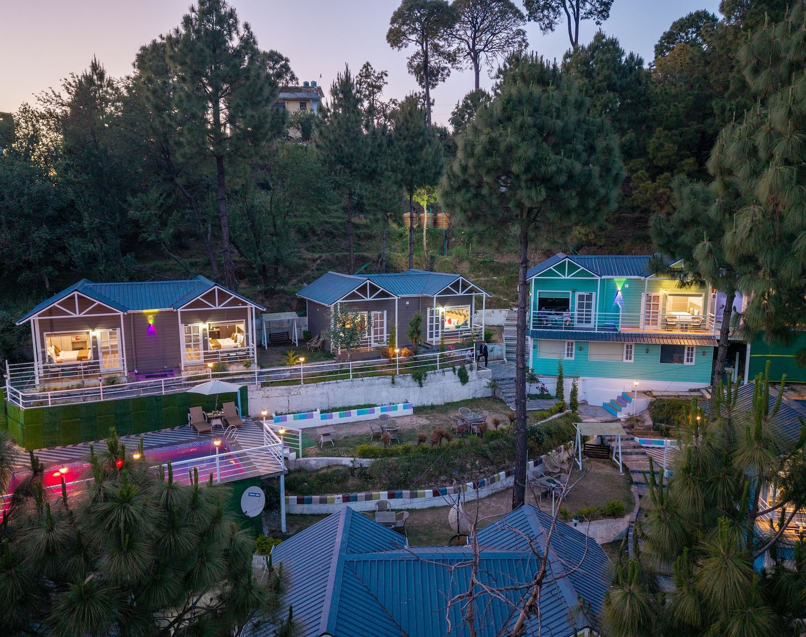 Several illuminated cabins are nestled on a hillside covered with pine trees at dawn - DLS Nature Trinket Resort, Dalhousie.