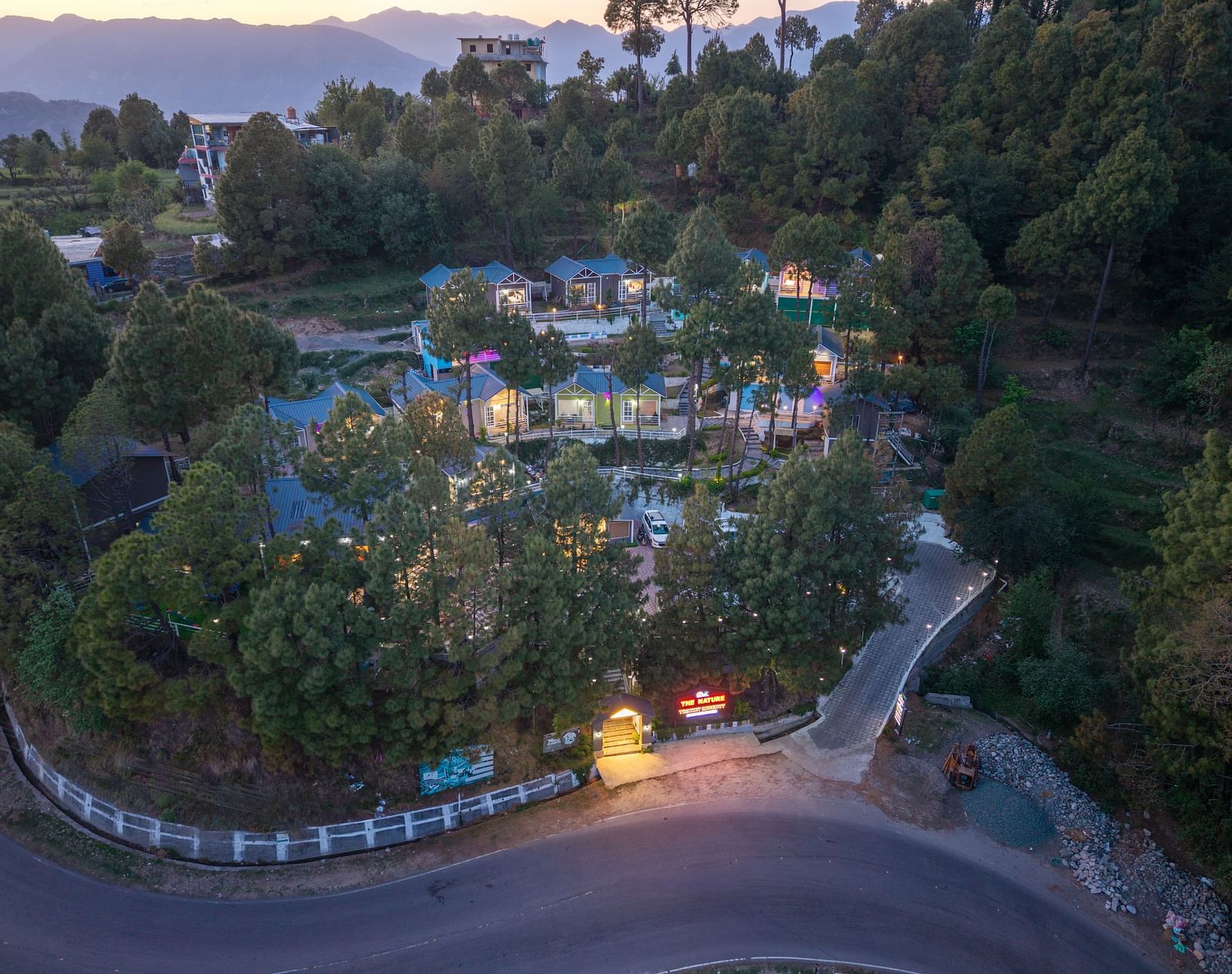 An aerial view shows a hillside resort with multiple buildings nestled among trees at dusk - DLS Nature Trinket Resort, Dalhousie.