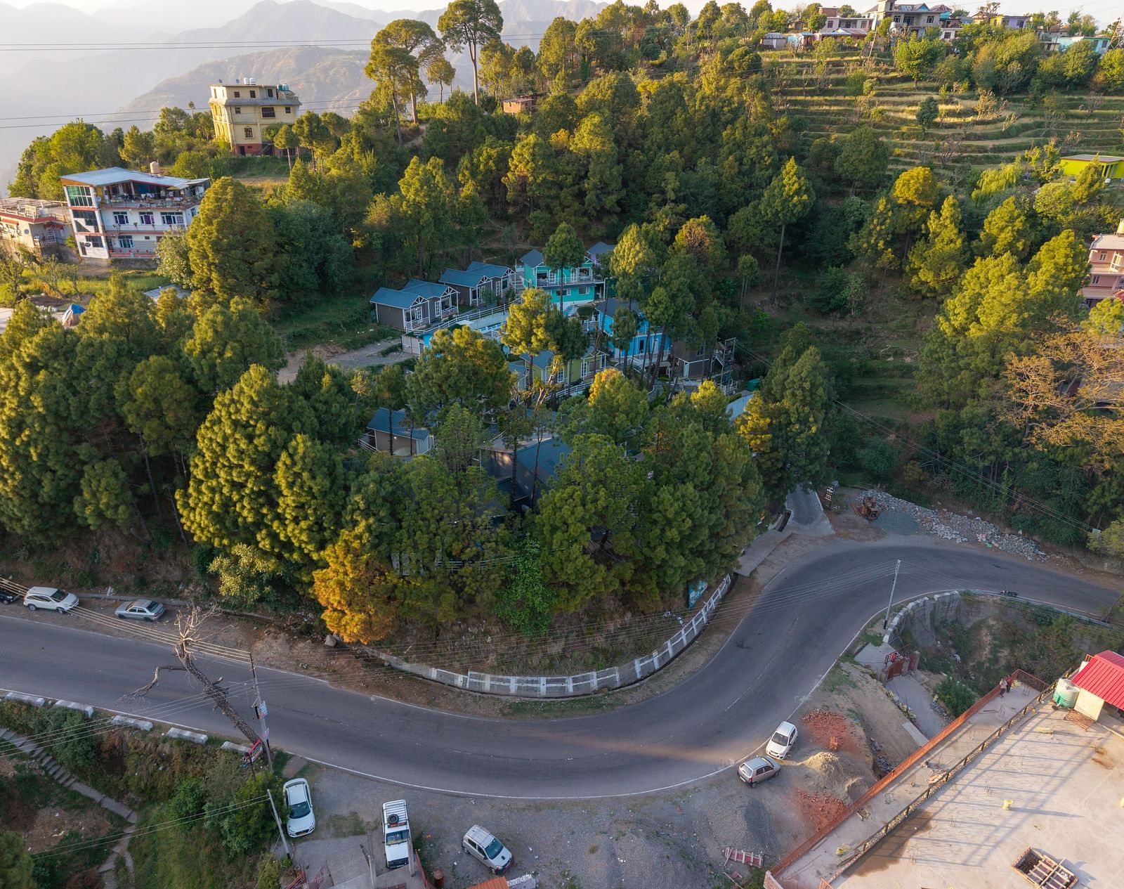 Aerial view of a hillside resort surrounded by dense greenery, located on a sharp bend in a mountain road with scattered buildings and terraced slopes in the background - DLS Nature Trinket Resort, Dalhousie.