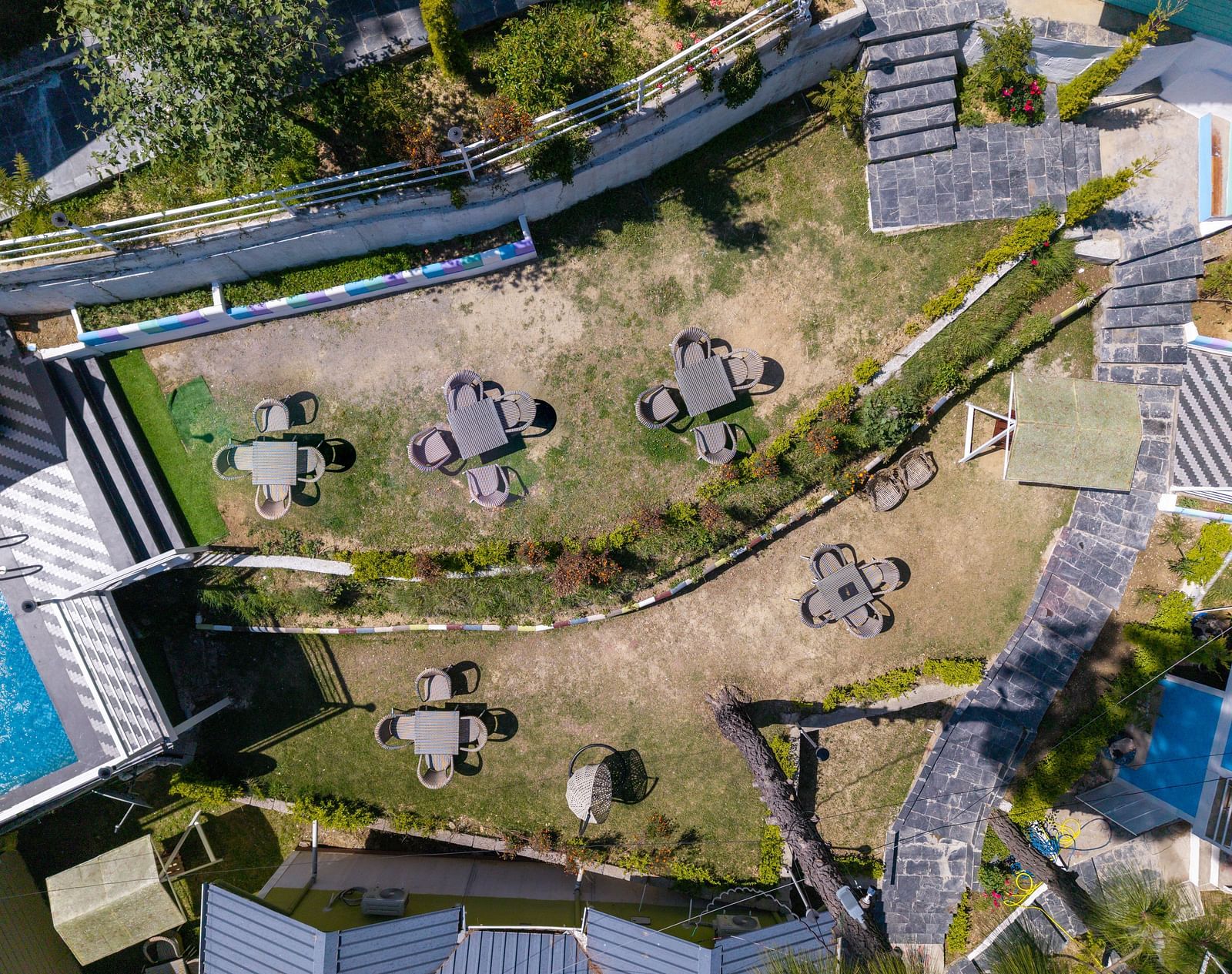Aerial view of a terraced garden area with multiple round tables and chairs arranged on grassy levels, surrounded by walkways, plants, and nearby buildings - DLS Nature Trinket Resort, Dalhousie.