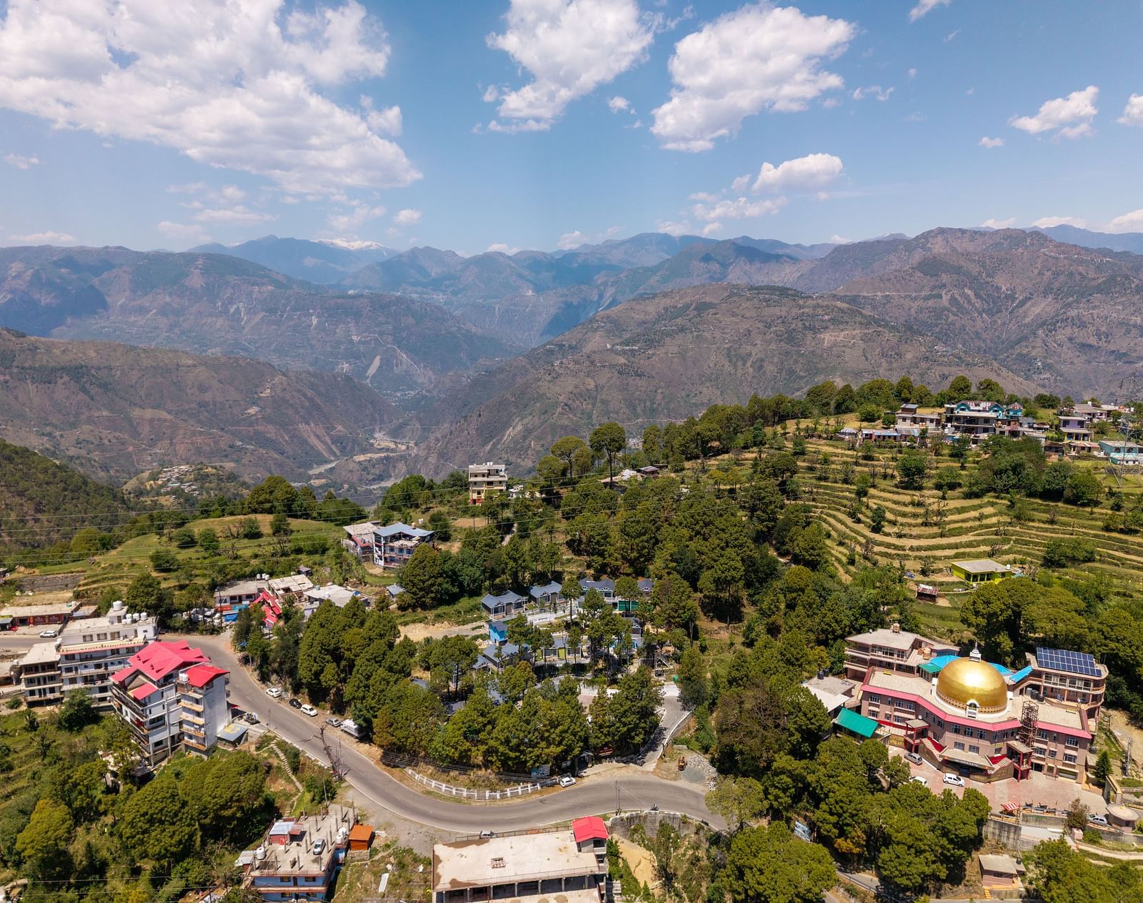 An aerial view of rolling hills, with residential and commercial buildings among lush greenery. A winding road connects the structures, and a temple with a golden dome stands out - DLS Nature Trinket Resort, Dalhousie.