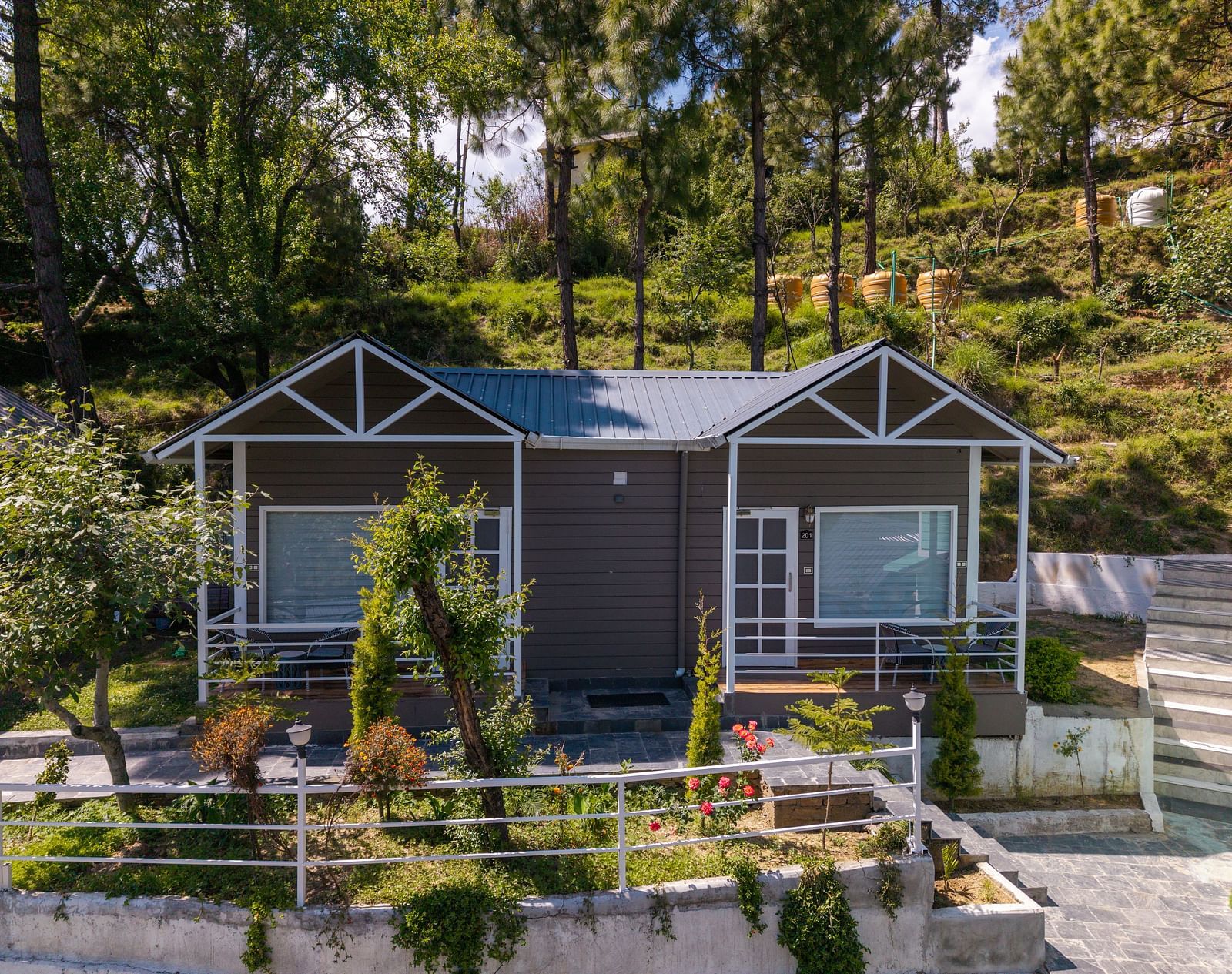Charming hillside cottage with a twin-gabled roof, large front windows, and a landscaped garden surrounded by trees and greenery - DLS Nature Trinket Resort, Dalhousie.