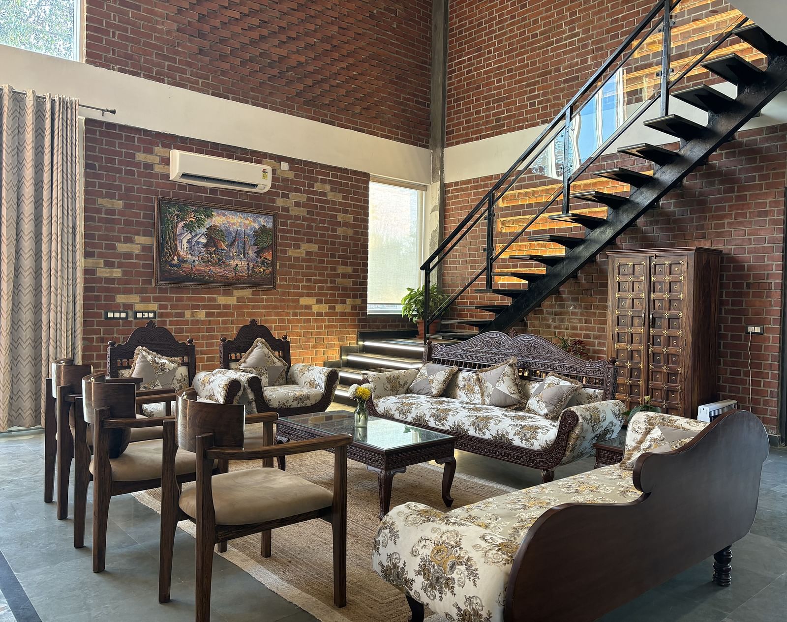 A living room at Tisya Farms, Gurgaon with multiple sofas, large windows, brick walls, and an overhead staircase.