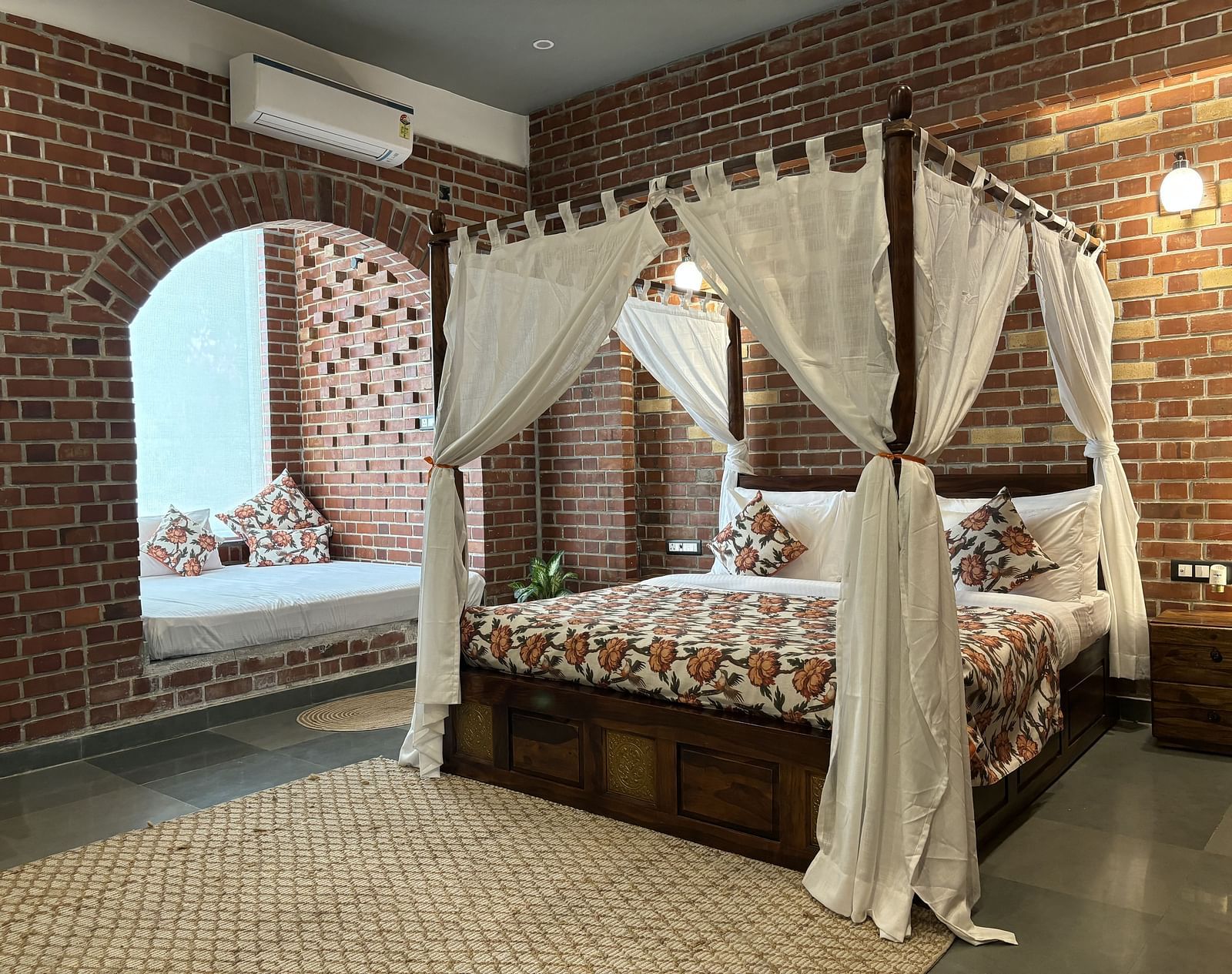 A bedroom at Tisya Farms, Gurgaon with a four-poster bed, white curtains, brick walls, and an arched window.