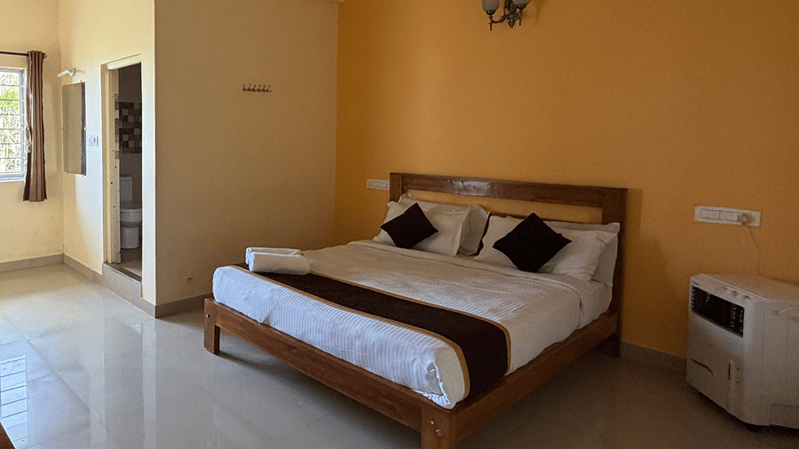 A spacious bedroom with a wooden double bed, a ceiling fan, and a wardrobe next to a small table with a red cloth | Kollanoor Guest House, Coorg