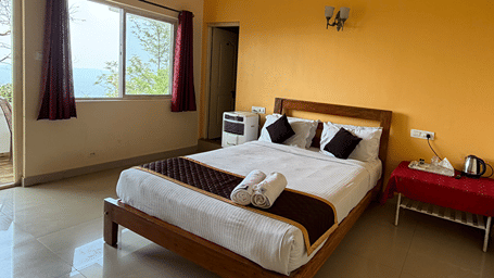 Double Room at Kollanoor Guest House with a wooden queen bed, a brown runner with gold trim, rolled towels, maroon curtains, and a hill-view window.