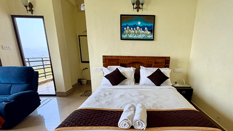 A wooden double bed with white sheets and brown runners, with a blue sofa visible to the left | Kollanoor Guest House, Coorg