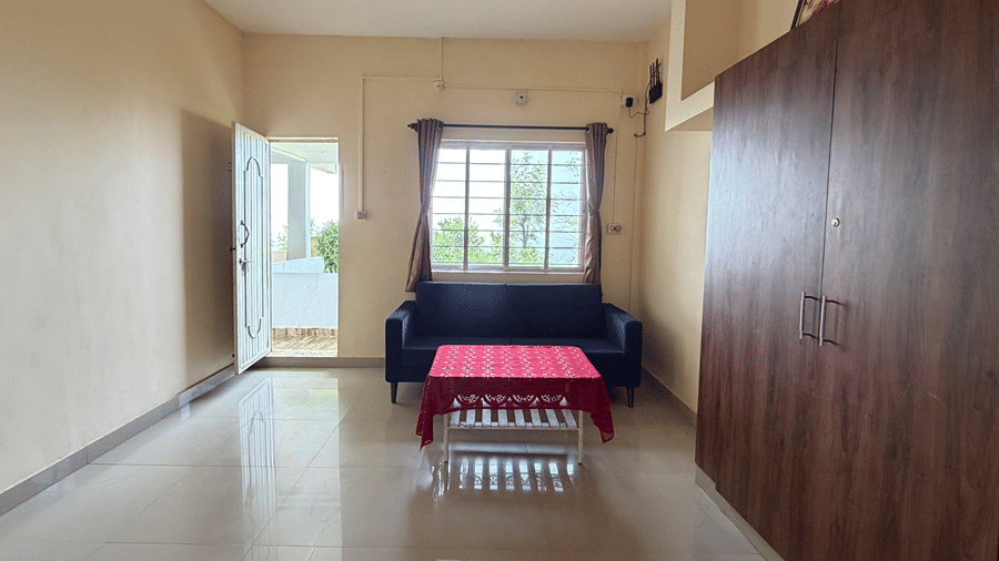 A large room with a dark sofa, a small table covered in a red cloth, and a tall wooden wardrobe along the wall | Kollanoor Guest House, Coorg