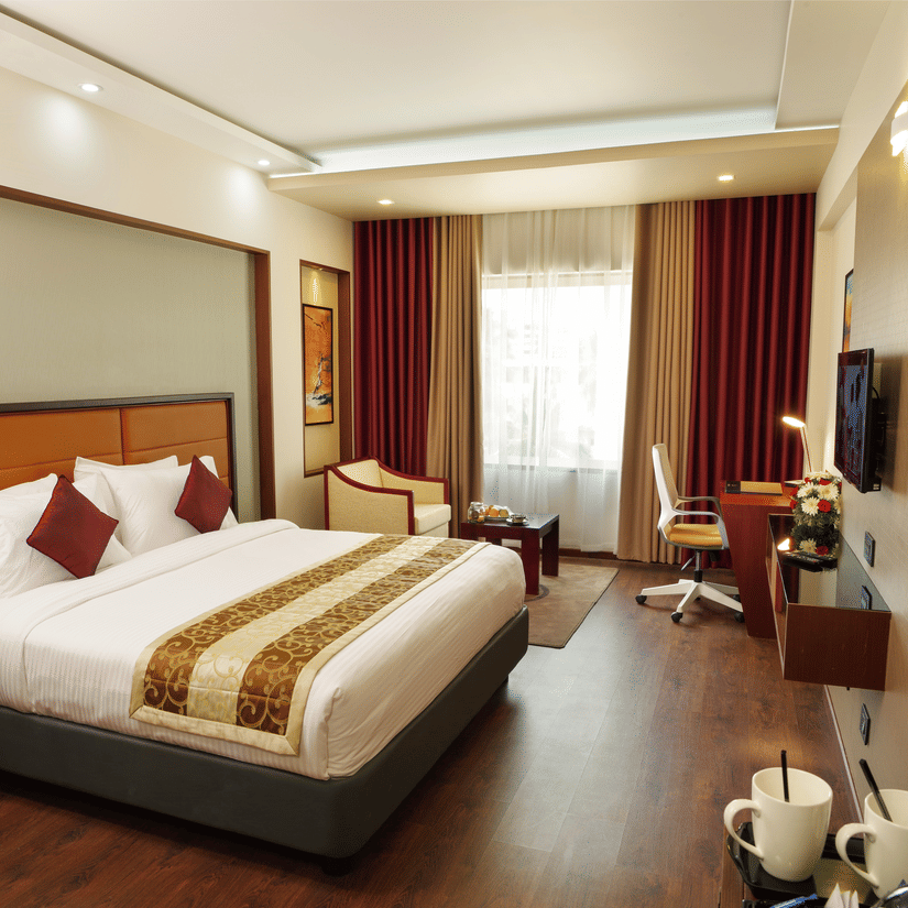 The Standard Room at Dimora, Thiruvananthapuram with a king size bed, a work desk, a flat screen television, a glass window with curtains, and wooden flooring.