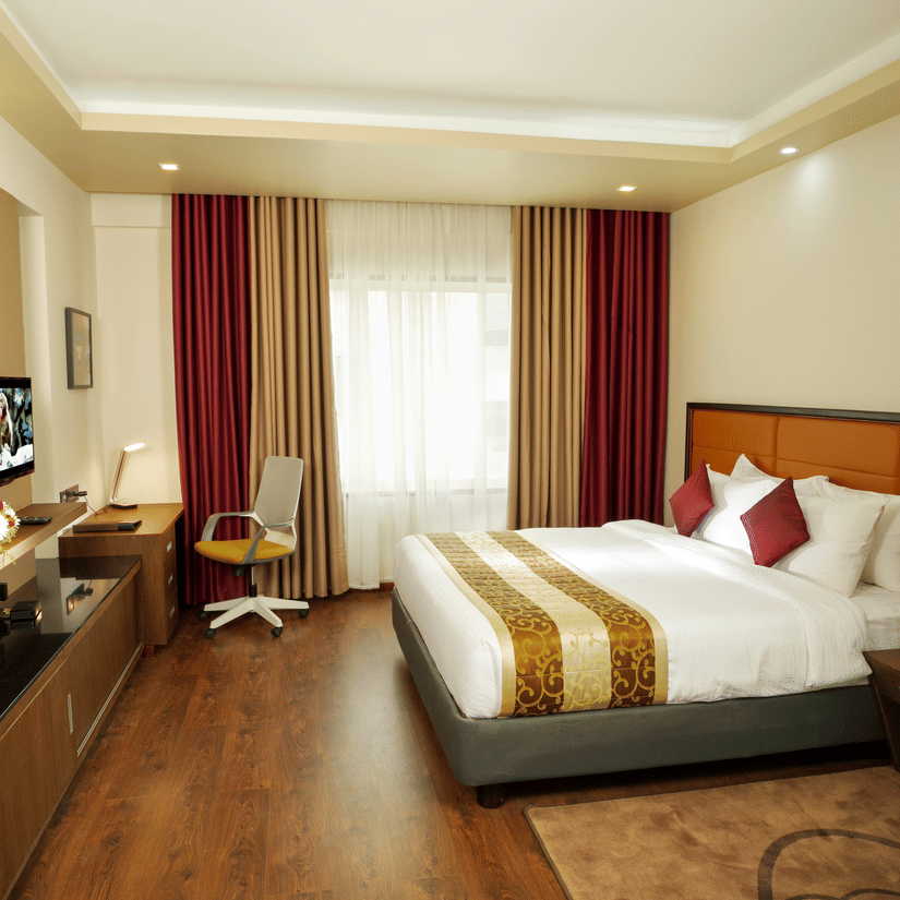 The Suite at Dimora, Thiruvananthapuram with a king size bed, a glass window with long curtains, ceiling lights, wooden flooring, and work desk.