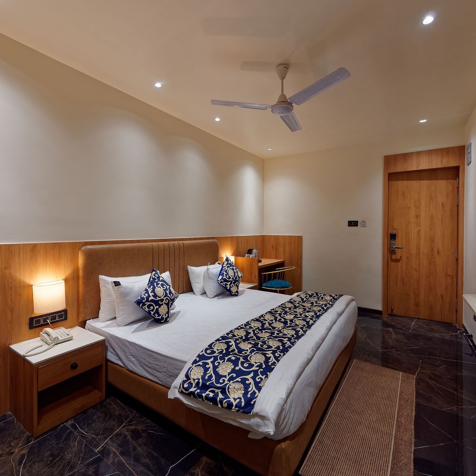 Image of a studio room with a bed with a table on the side of the bed and a television in front Image of a bed with table and chair on the side of the table and a fan on te ceiling at Ankit Hotel, Jabalpur