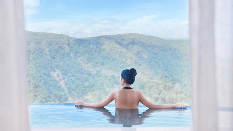 Woman relaxes in an infinity pool at Polo Resort Cherrapunji, leaning on the edge while gazing at forested rolling hills under a partly cloudy blue sky.