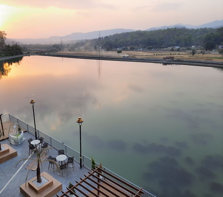 An outside pool area view of a lake surrounded by hills, with a seating area and a walkway along the shore, during the sunset  time.